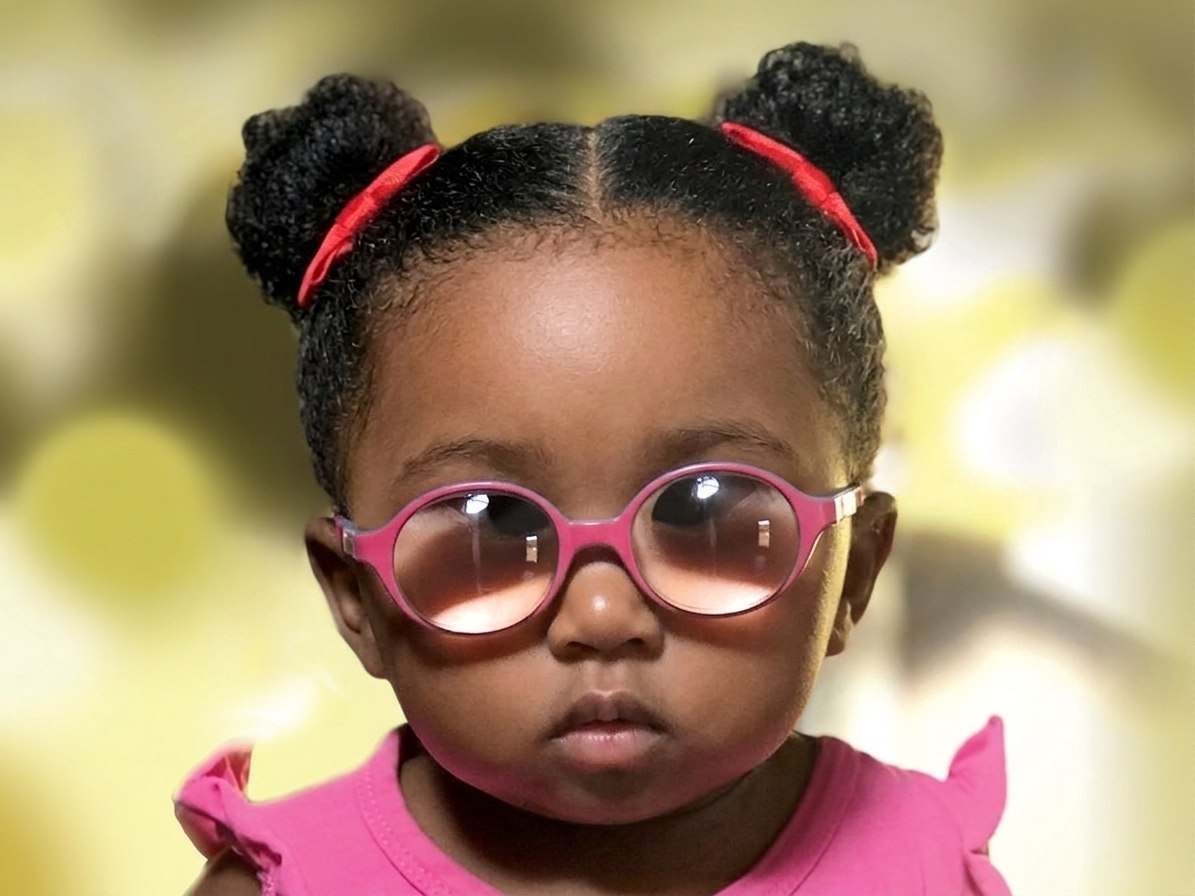A toddler wears round pink glasses, red bows in her hair, gazing steadily at the camera.