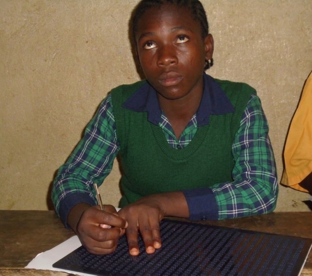 A teenage student rests her fingers on a braille board at a wooden school desk, pen in hand.